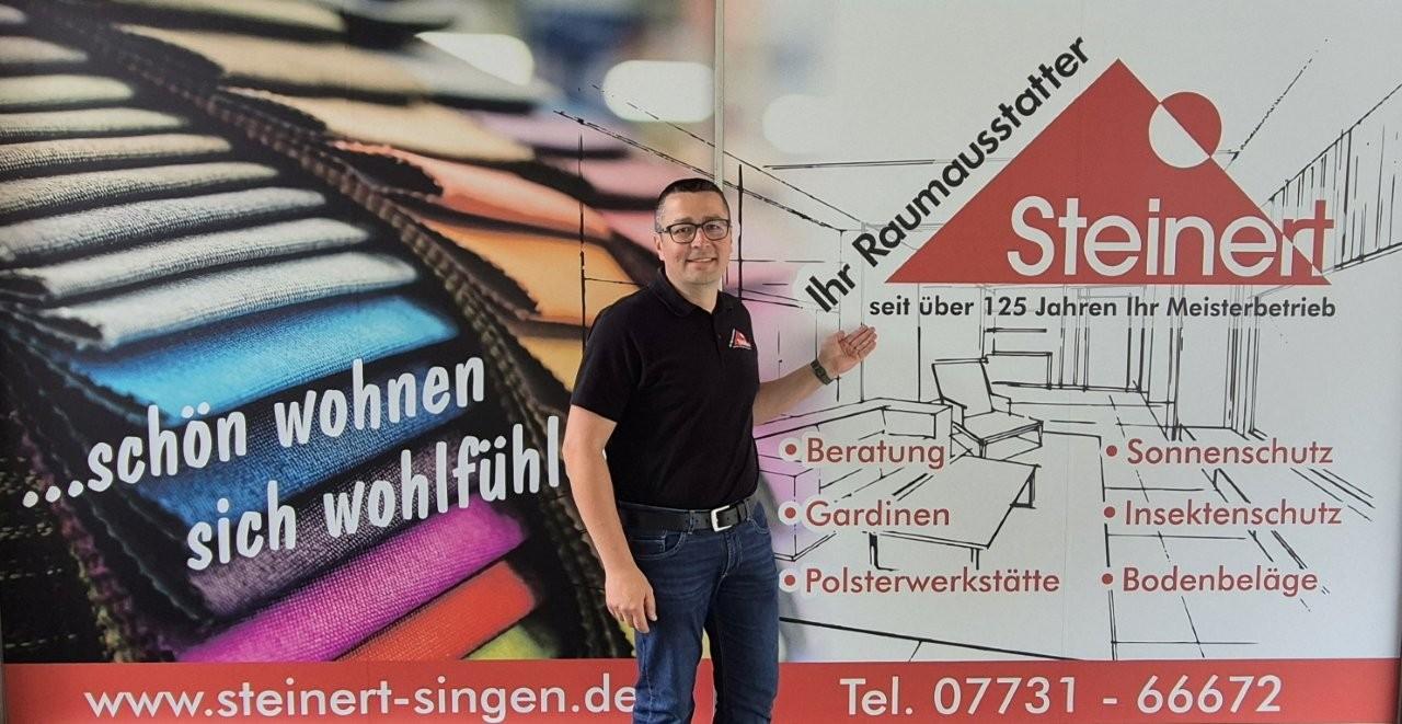 Herr Steinert vor Werbebanner mit bunten Stoffmustern und Firmenlogo 'Steinert'